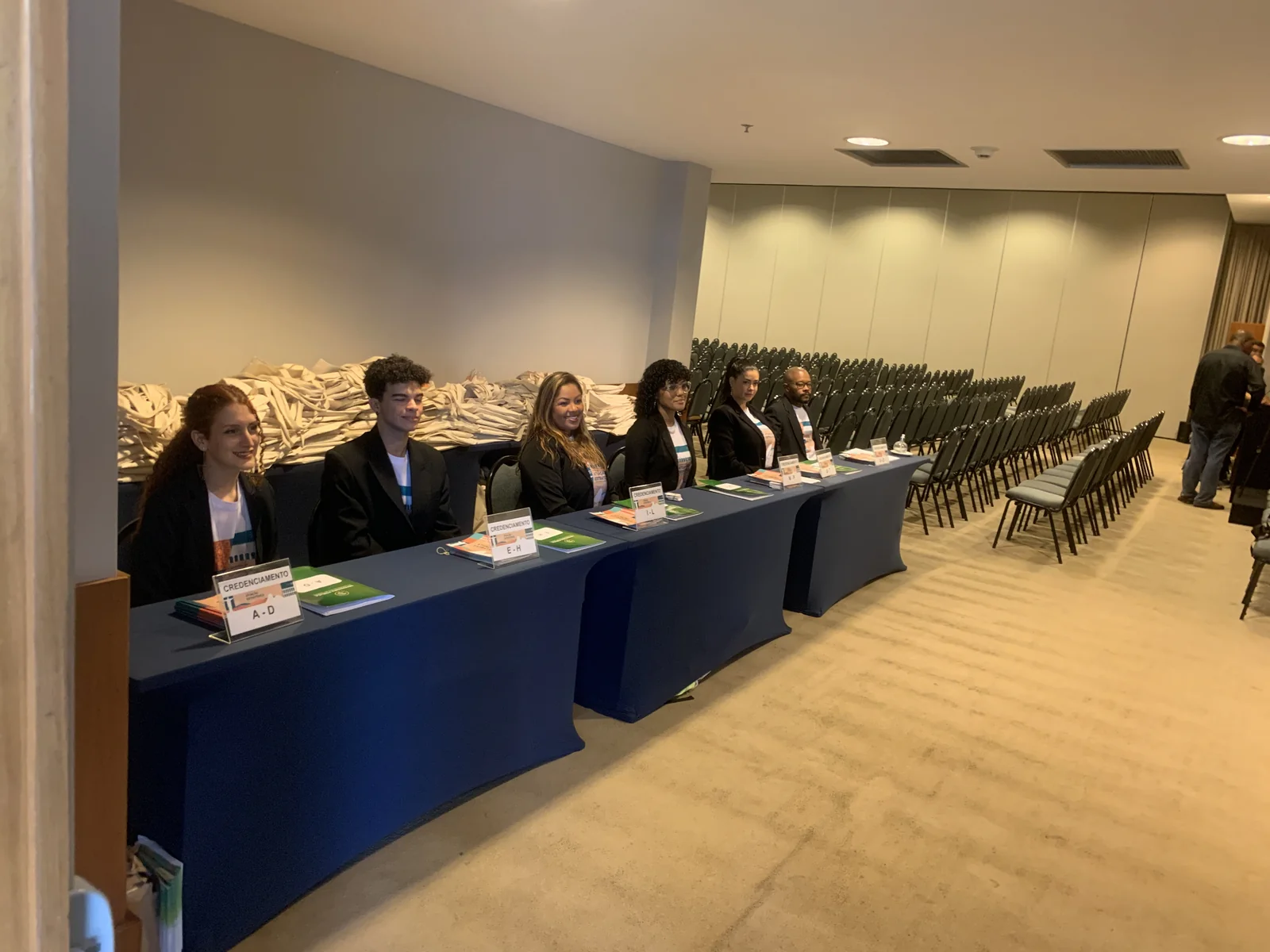 Mesa de credenciamento com a equipe de recepção sentada, ao lado das cadeiras do auditório.