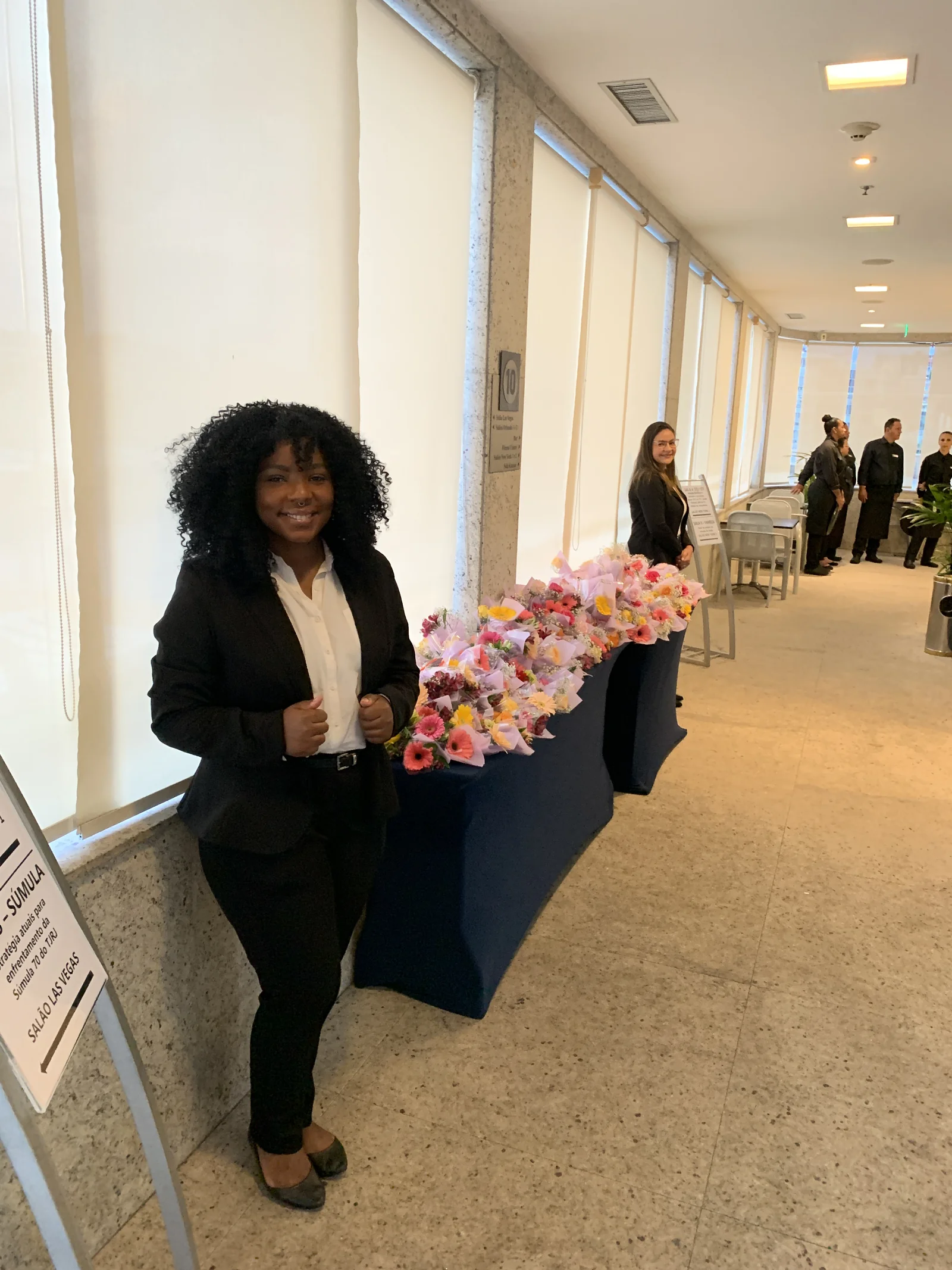 Recepcionista ao lado de uma mesa com arranjos de flores no corredor do evento.