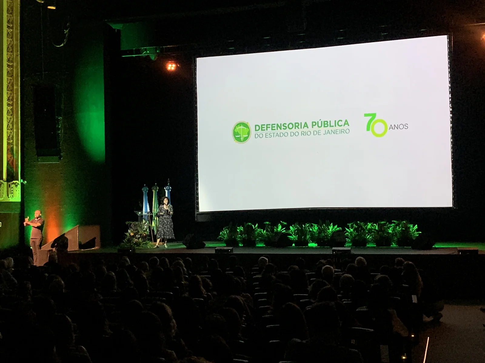 Telão com a marca dos 70 anos da Defensoria Pública no palco do Teatro Riachuelo, diante da plateia.