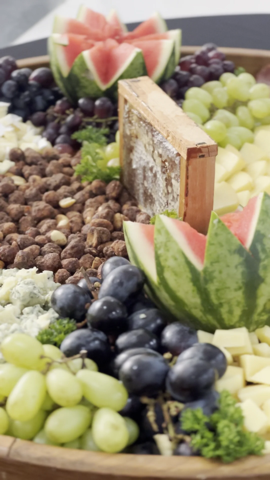 Tábua de madeira pronta com favo de mel, uvas verdes e roxas, queijos, castanhas e melancia esculpida.