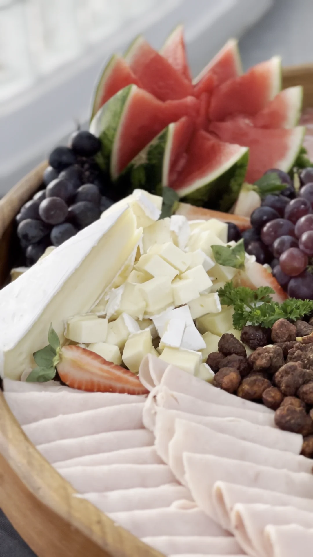 Detalhe de tábua de frios pronta com queijo brie, cubos de queijo, peito de peru, uvas, morangos e melancia esculpida.