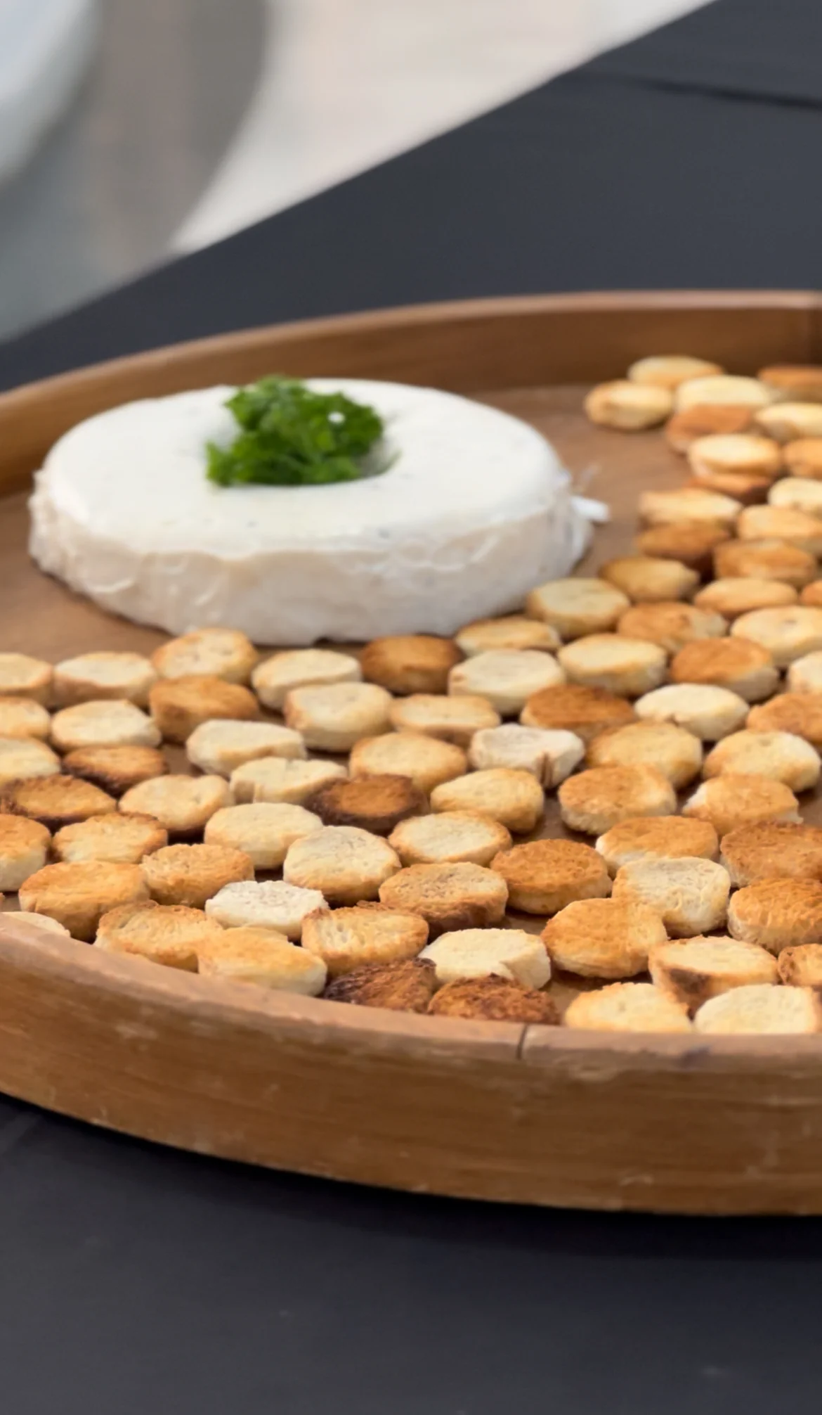 Tábua de madeira redonda com queijo cremoso decorado e torradas organizadas em espiral sobre toalha preta.
