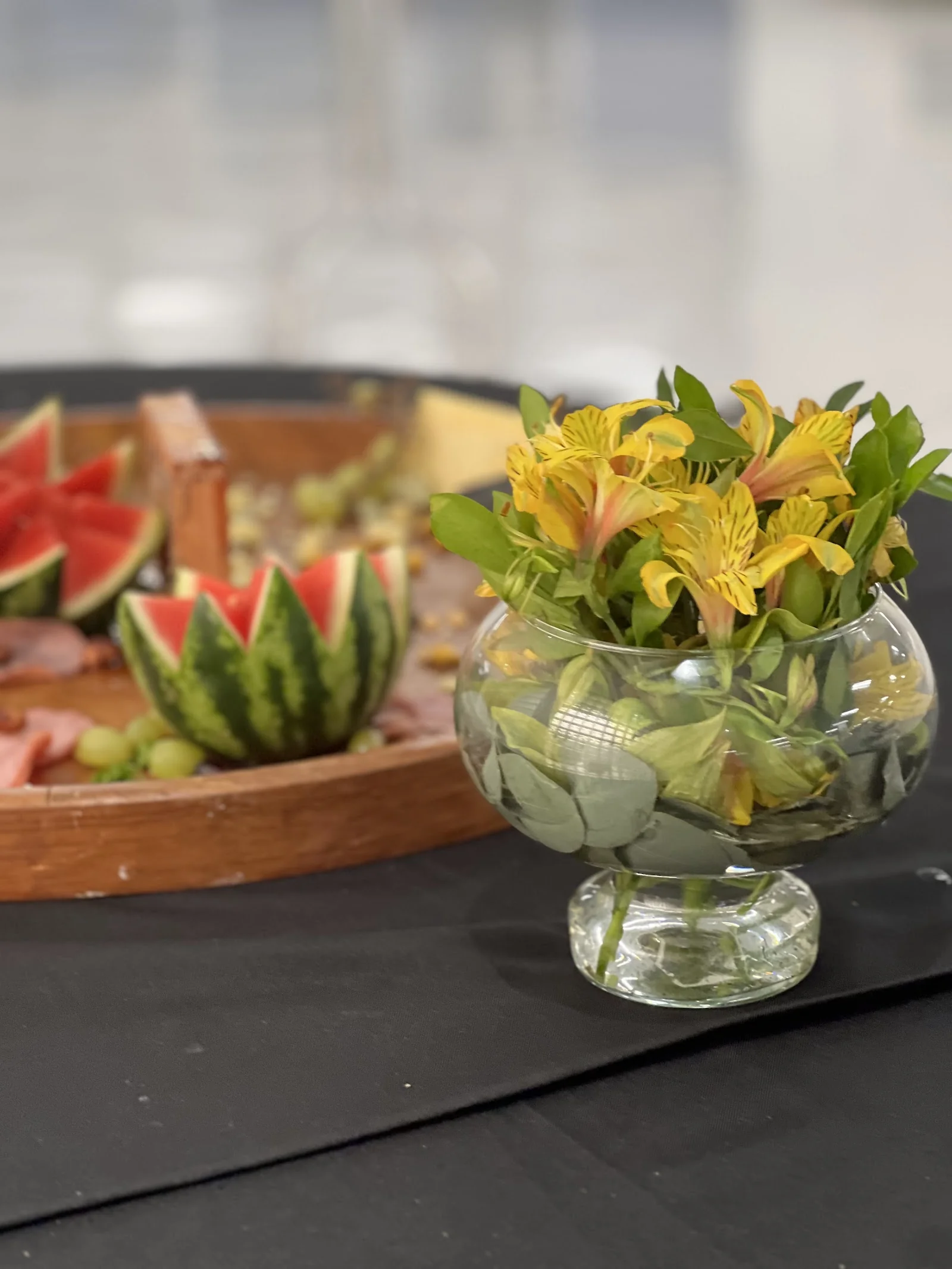 Arranjo floral amarelo em vaso de vidro sobre mesa com toalha preta e tábua de frutas e favo de mel ao fundo.