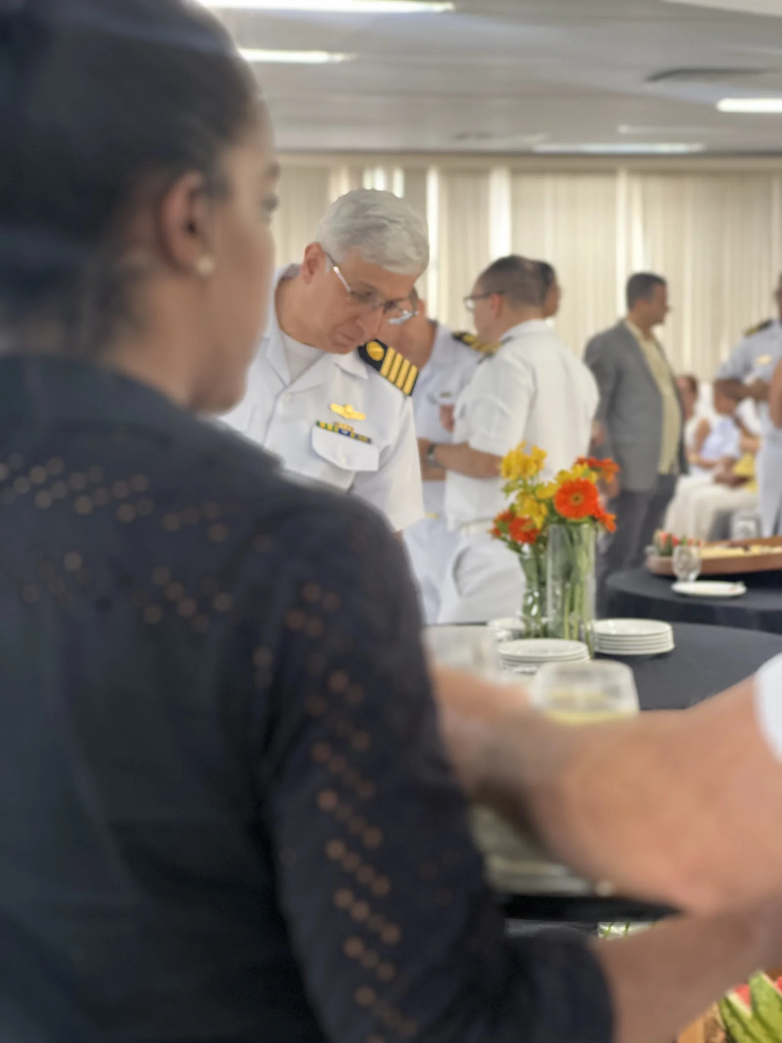 Oficiais da Marinha conversam no salão da Escola de Guerra Naval ao lado das mesas decoradas com arranjos florais.