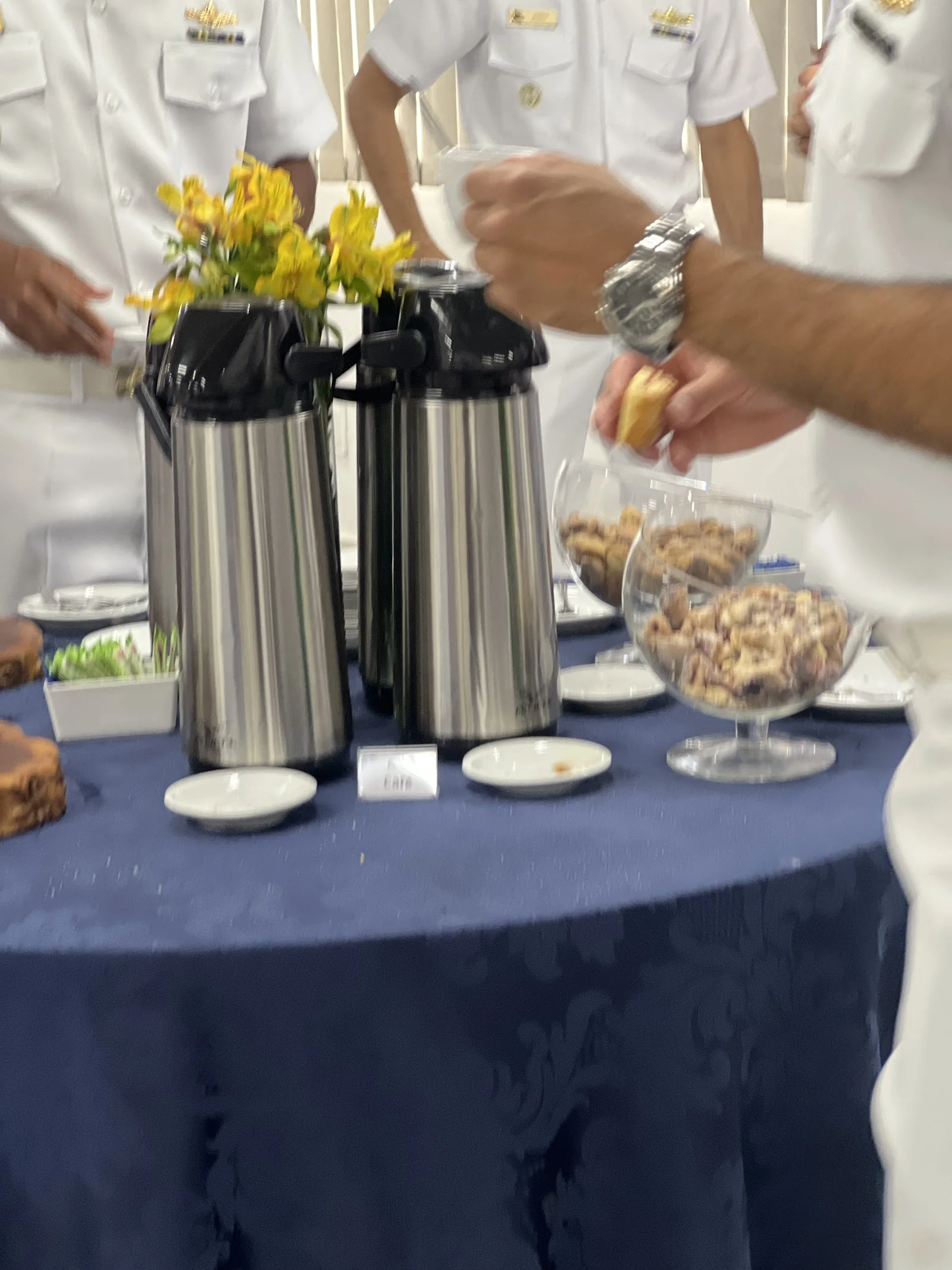 Oficiais da Marinha em uniforme branco servem-se de café e biscoitos na mesa com toalha azul.