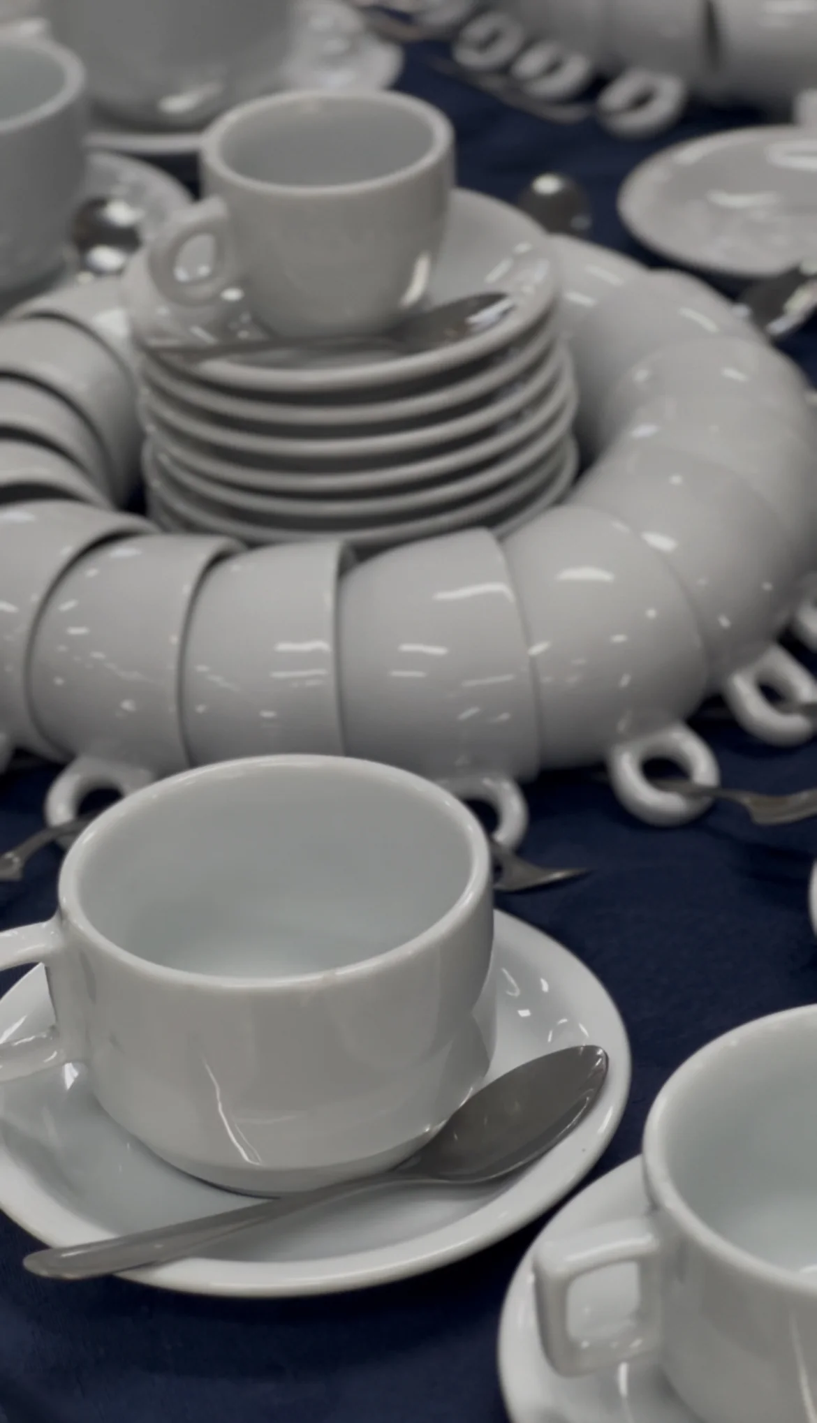 Xícaras e pires de porcelana branca organizados em roda na mesa de café sobre toalha azul.